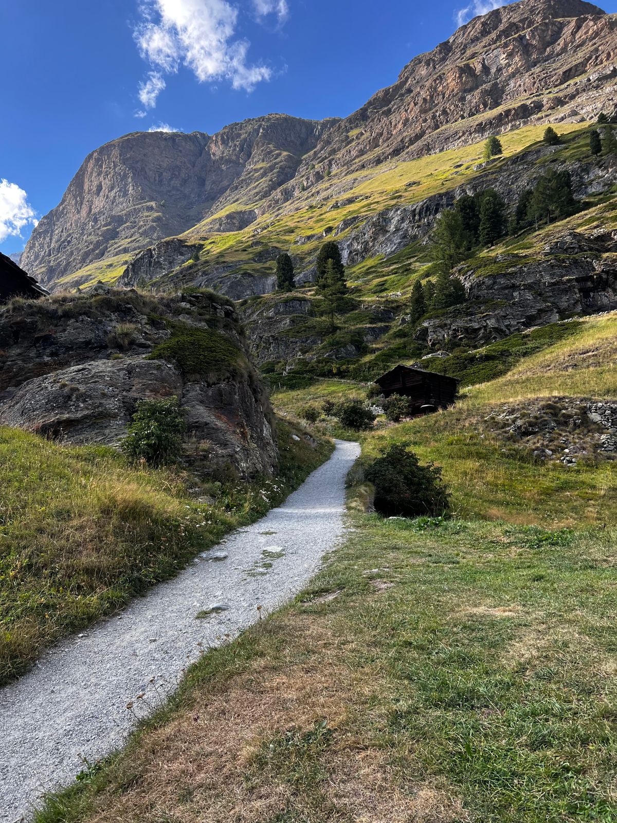 Chemin de terre traversant une prairie de fond de vallée, au pied d’une paroi rocheuse